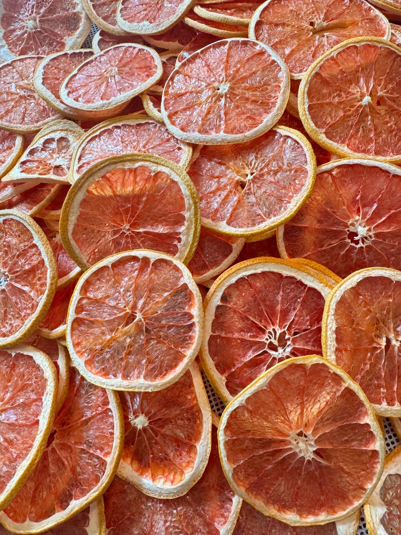 May include: A close-up shot of numerous dried grapefruit slices. The slices are a vibrant reddish-orange color with a pale yellow rind. The arrangement is dense, showcasing the texture and detail of the dehydrated fruit, perfect for culinary or decorative use.