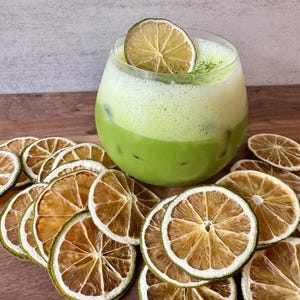 May include: A refreshing green matcha drink in a clear glass, garnished with a dried lime slice. Surrounding the drink are several dried lime slices, arranged on a wooden surface. The drink has a layered appearance with ice visible.