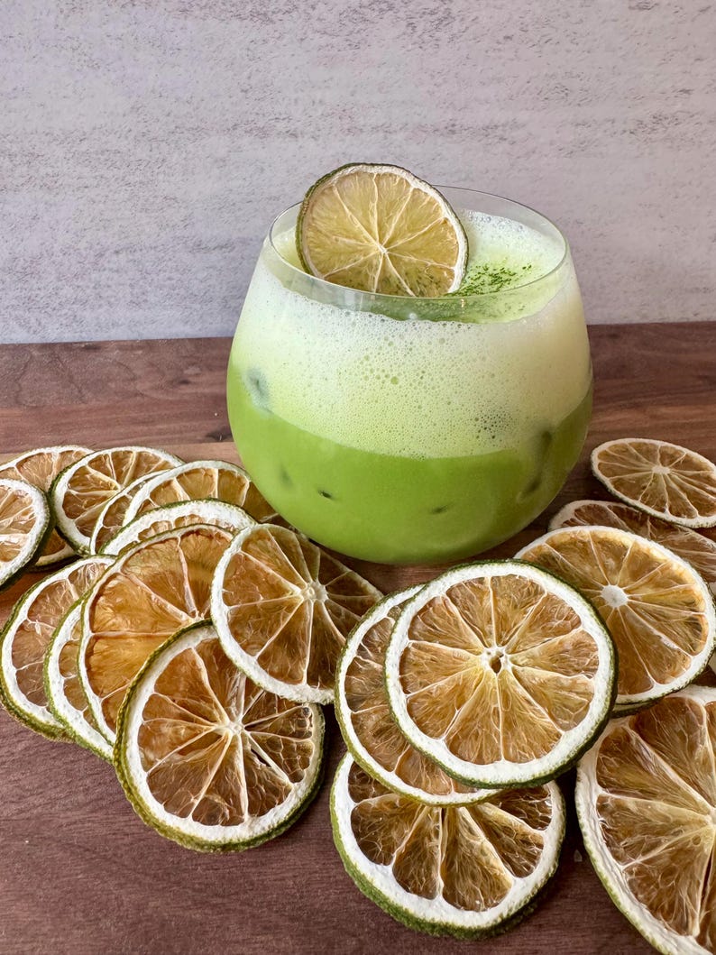May include: A refreshing green matcha drink in a clear glass, garnished with a dried lime slice. Surrounding the drink are several dried lime slices, arranged on a wooden surface. The drink has a layered appearance with ice visible.