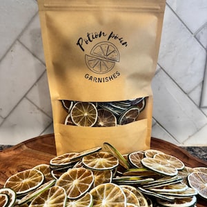May include: A brown paper bag labeled "Potion Pour Garnishes" filled with dried lime slices, with more slices scattered in front. The bag has a clear window showing the contents. The lime slices are circular and have a brown and green color.