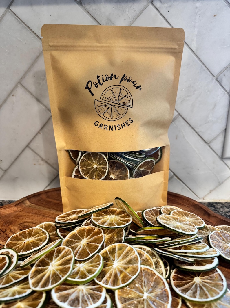 May include: A brown paper bag labeled "Potion Pour Garnishes" filled with dried lime slices, with more slices scattered in front. The bag has a clear window showing the contents. The lime slices are circular and have a brown and green color.