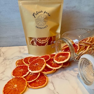 May include: A kraft paper bag and a glass jar filled with dried orange slices. The bag has a clear window and the text "Potion pour GARNISHES" printed on it. Orange slices are scattered on a marble surface.