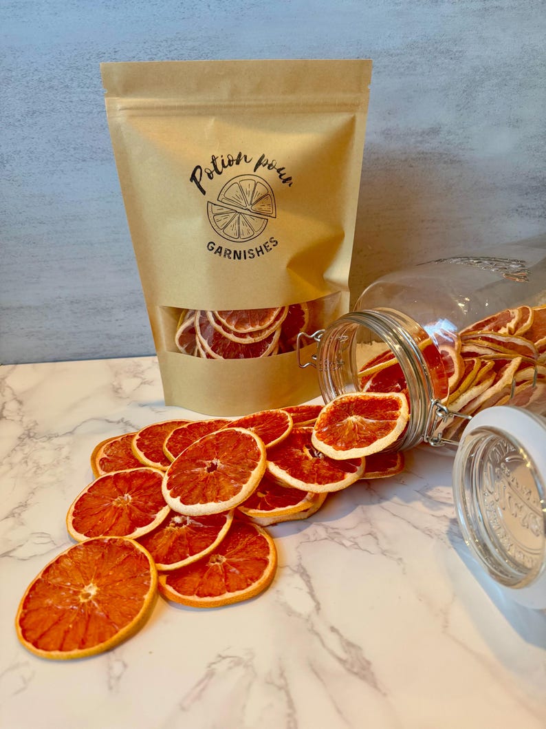 May include: A kraft paper bag and a glass jar filled with dried orange slices. The bag has a clear window and the text "Potion pour GARNISHES" printed on it. Orange slices are scattered on a marble surface.