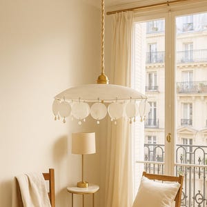 Op de afbeelding: Een witte hanglamp met een ronde kap en schelpachtige accenten hangt aan een gedraaid gouden snoer. De lamp staat in een kamer met een raam, stoelen en een kleine tafellamp.