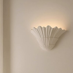 Könnte beinhalten: Eine weiße Wandleuchte mit einem strukturierten, muschelartigen Design. Die Leuchte hat einen gewellten Rand und strahlt ein warmes, diffuses Licht aus. Sie ist an einer neutralfarbenen Wand montiert.