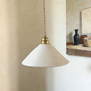 Può includere: Una lampada a sospensione bianca a forma di cono con un raccordo in ottone e un cavo marrone rivestito in tessuto. La luce pende dal soffitto. Lo sfondo include un dipinto con una natura morta.