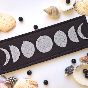 May include: Black fabric patch with white embroidered moon phases. The patch features a crescent moon, half moon, gibbous moon, full moon, and waning gibbous moon.