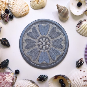 Könnte beinhalten: Ein kreisförmiger, hellblauer, gestickter Aufnäher mit einem Blumendesign, umgeben von verschiedenen Muscheln und kleinen schwarzen Perlen auf einer weißen Oberfläche. Der Aufnäher hat ein detailliertes, symmetrisches Blumenmuster.