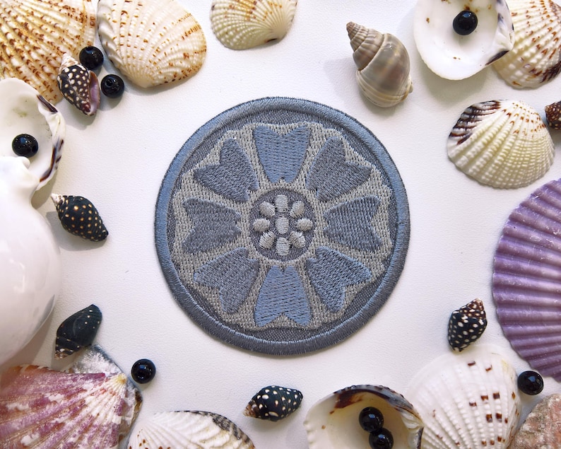May include: A circular, light blue embroidered patch with a floral design, surrounded by various seashells and small black beads on a white surface. The patch features a detailed, symmetrical flower pattern.