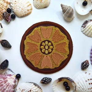 May include: Embroidered patch featuring a stylized flower design in shades of gold and brown, set against a brown circular border. The patch is surrounded by seashells and small black beads on a white surface, creating a decorative arrangement.