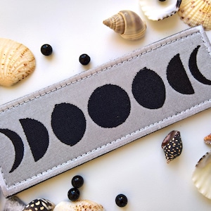May include: A gray fabric patch with black embroidered moon phases. The patch has a white border and is stitched with a white thread.