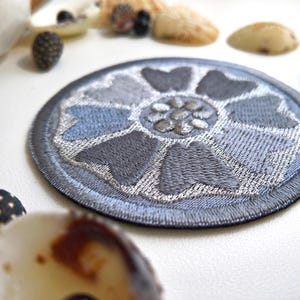 May include: A round, embroidered patch featuring a floral design in shades of gray and blue. The patch has a dark gray border and a detailed, textured appearance. Seashells and other natural elements are in the background.