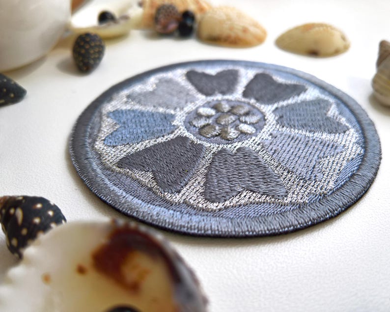 May include: A round, embroidered patch featuring a floral design in shades of gray and blue. The patch has a dark gray border and a detailed, textured appearance. Seashells and other natural elements are in the background.