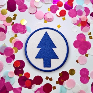 May include: A circular embroidered patch featuring a solid blue pine tree icon centred on a white background with a thick blue stitched border. The patch is surrounded by a scattered arrangement of pink, magenta, and gold circular confetti.