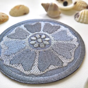 May include: A round, embroidered patch with a floral design in shades of gray and silver. The patch features a detailed, textured pattern and a dark gray border. Seashells are scattered around the patch.