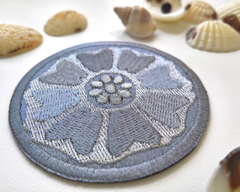 May include: A round, embroidered patch with a floral design in shades of gray and silver. The patch features a detailed, textured pattern and a dark gray border. Seashells are scattered around the patch.