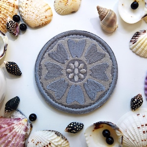 Puede incluir: Un parche redondo y gris bordado con un diseño floral, rodeado de conchas marinas y cuentas negras sobre una superficie blanca. El parche presenta un patrón floral azul claro y un borde gris.