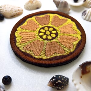 K&ouml;nnte beinhalten: Ein runder, gestickter Aufn&auml;her mit floralem Design. Der Aufn&auml;her hat eine gelbe Mitte mit braunen Bl&uuml;tenblattformen und einen braunen Rand. Muscheln und kleine schwarze Perlen umgeben den Aufn&auml;her.