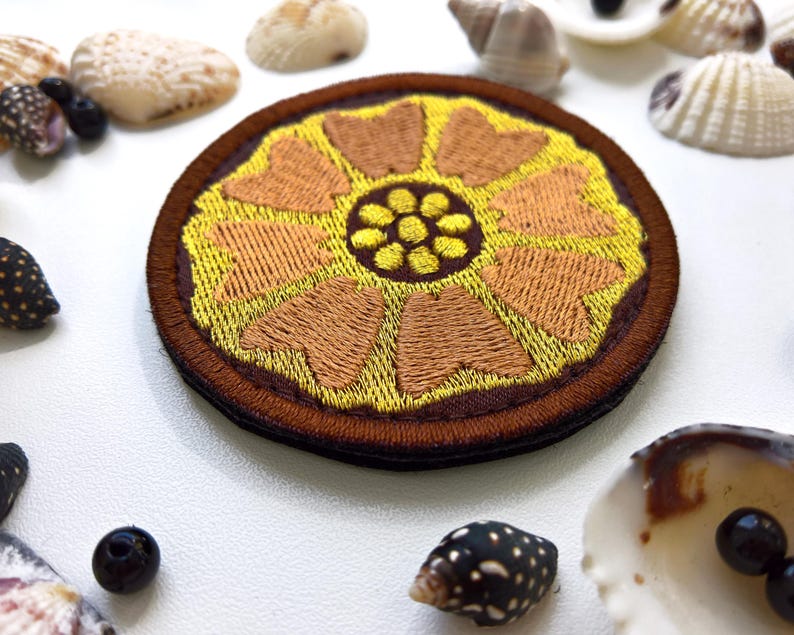 K&ouml;nnte beinhalten: Ein runder, gestickter Aufn&auml;her mit floralem Design. Der Aufn&auml;her hat eine gelbe Mitte mit braunen Bl&uuml;tenblattformen und einen braunen Rand. Muscheln und kleine schwarze Perlen umgeben den Aufn&auml;her.