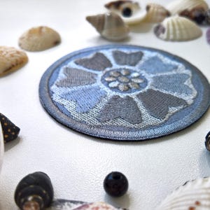 May include: A round, embroidered patch featuring a stylized blue and gray lotus flower design. The patch is surrounded by a dark blue border. The image also includes seashells and small, dark-colored beads.