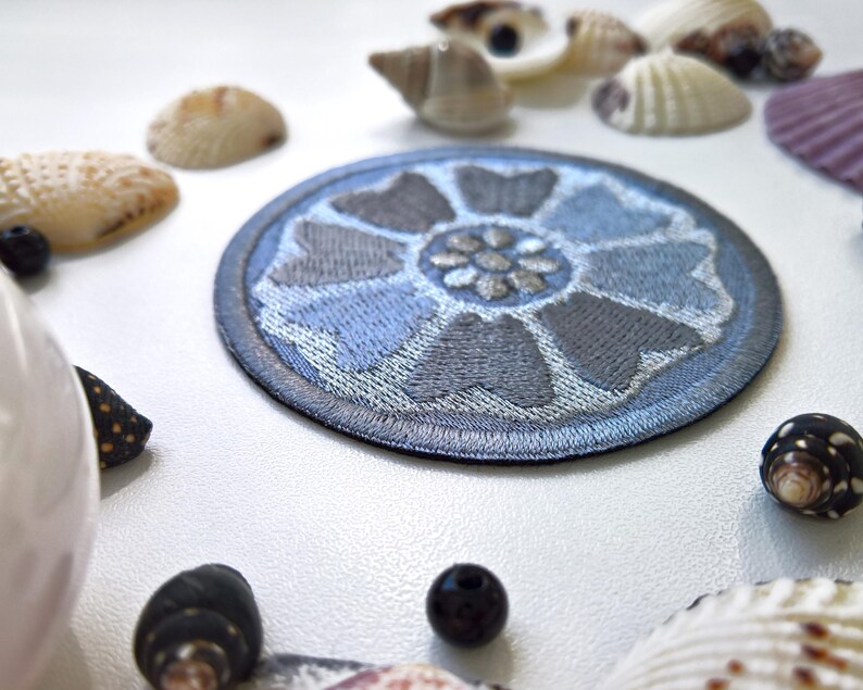 May include: A round, embroidered patch featuring a stylized blue and gray lotus flower design. The patch is surrounded by a dark blue border. The image also includes seashells and small, dark-colored beads.