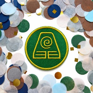 Può includere: Una toppa ricamata circolare con sfondo verde scuro, bordo giallo e un simbolo centrale giallo che raffigura una spirale sopra due linee orizzontali, circondata da una collezione sparsa di cerchi di coriandoli di carta multicolori.