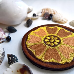 K&ouml;nnte beinhalten: Ein runder, gestickter Aufn&auml;her mit einem floralen Design in Gold-, Braun- und Oranget&ouml;nen. Der Aufn&auml;her ist von einem braunen Rand umgeben und wird zwischen Muscheln und kleinen dekorativen Elementen platziert, was ein Strandthema erzeugt.