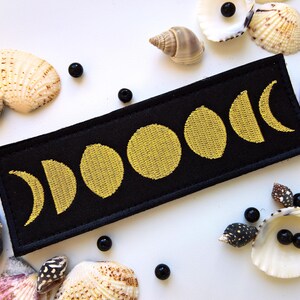 Könnte beinhalten: Ein schwarzer Stoff-Patch mit einem goldenen gestickten Design der Mondphasen. Der Patch ist von Muscheln und Perlen umgeben.