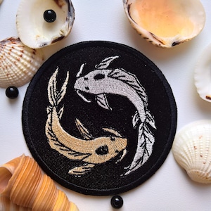 Puede incluir: Un parche bordado negro con dos peces koi nadando en direcciones opuestas. Los peces son blancos y dorados con contornos negros. El parche es circular y tiene un fondo negro.