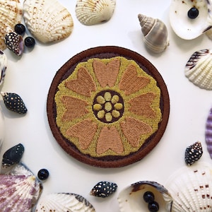 May include: Embroidered patch featuring a stylized flower design in shades of gold and brown, surrounded by a brown border. The patch is set against a white background, adorned with various seashells and small black beads, creating a coastal theme.