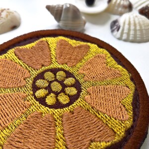 K&ouml;nnte beinhalten: Runder, gestickter Aufn&auml;her mit einem floralen Design in Gold-, Braun- und Gelbt&ouml;nen. Der Aufn&auml;her hat ein detailliertes, strukturiertes Muster. Muscheln sind im Hintergrund. Geeignet f&uuml;r Kleidung oder Accessoires, zum Aufn&auml;hen oder Aufb&uuml;geln.