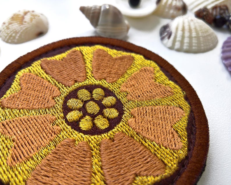 K&ouml;nnte beinhalten: Runder, gestickter Aufn&auml;her mit einem floralen Design in Gold-, Braun- und Gelbt&ouml;nen. Der Aufn&auml;her hat ein detailliertes, strukturiertes Muster. Muscheln sind im Hintergrund. Geeignet f&uuml;r Kleidung oder Accessoires, zum Aufn&auml;hen oder Aufb&uuml;geln.