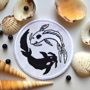 Puede incluir: Símbolo yin yang bordado en blanco y negro con dos peces, uno blanco y otro negro, nadando en direcciones opuestas. El símbolo está sobre un fondo blanco y está rodeado de conchas.