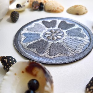 May include: A round, embroidered patch with a floral design in shades of gray and blue. The patch is surrounded by seashells and small black beads, creating a coastal theme. The patch is likely for clothing or accessories.