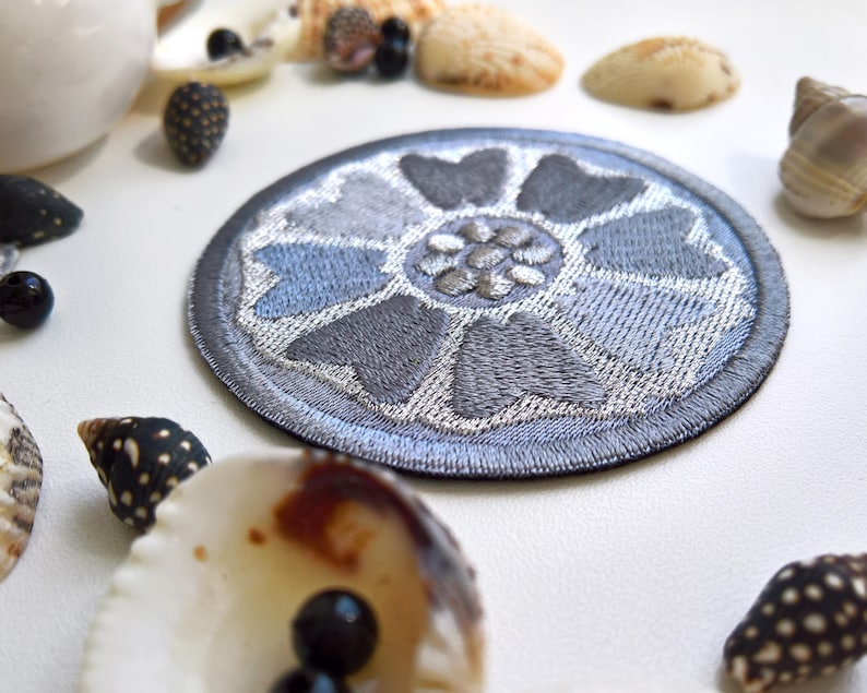 May include: A round, embroidered patch with a floral design in shades of gray and blue. The patch is surrounded by seashells and small black beads, creating a coastal theme. The patch is likely for clothing or accessories.