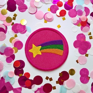 May include: A circular embroidered patch featuring a bright yellow shooting star with a trailing rainbow tail consisting of purple, green, and orange stripes, all set against a solid hot pink background with a matching hot pink stitched border.