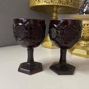 Op de afbeelding: Twee robijnrode vintage kelken met sierlijke, getextureerde ontwerpen. De glazen hebben een ruitpatroon en staan op een zeshoekige basis. Het glaswerk is waarschijnlijk van glas en geschikt voor het serveren van wijn of andere dranken.