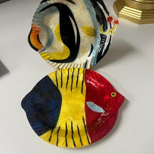 May include: Two decorative glass plates shaped like fish. The top plate features a design with black, white, yellow, and orange accents. The bottom plate has a red head, yellow body, and blue accents. Both plates have a glossy finish.