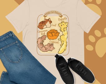 May include: A beige t-shirt with a graphic of five cats in different poses, each labeled with a bakery-themed name: Baguette, Loaf, Cinnamon Roll, Croissant, and a fifth cat with no label. The graphic is surrounded by a brown frame with the text "Kitty Cat Bakery" at the top.