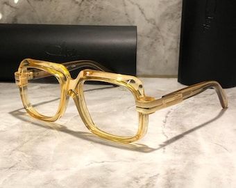 May include: A pair of eyeglasses with a light amber-colored frame and temples. The rectangular frames have a thick, bold design. The lenses are clear. A black case is in the background. The brand name is visible on the frame.