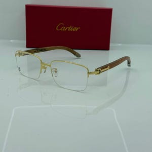 Op de afbeelding: Goudkleurige bril met rechthoekige glazen en houten pootjes. De bril wordt gepresenteerd voor een rode doos met de merknaam "Cartier" erop gedrukt. De bril heeft een reflecterend oppervlak.