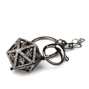 Pode incluir: Um porta-chaves em metal escuro com um pendente icosaedro oco em forma de gaiola, com motivos intrincados de caveiras incrustados na sua estrutura triangular. O pendente está preso a uma argola circular e a um fecho mosquetão a condizer.