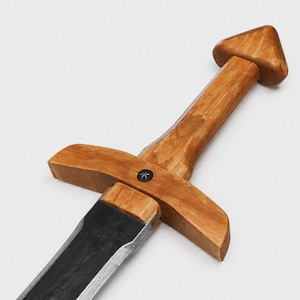 Könnte beinhalten: Ein Spielzeugschwert mit einem glatten, hellbraunen Holzgriff und einer Parierstange mit dreieckigem Knauf. Eine kleine, dunkle, kreisförmige Einlage mit sternförmigem Design befindet sich mittig auf der Parierstange. Die Klinge ist dunkelgrau.