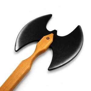Könnte beinhalten: Eine Doppelaxt mit einem großen, mattschwarzen Kopf und zwei geschwungenen Klingen. Der Kopf ist fest an einem hellen, glatten Holzstiel befestigt. In der Mitte befindet sich ein kleiner, kupferfarbener Metallniet.