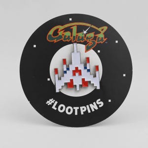 Könnte beinhalten: Eine weiß, rot und blau emaillierte Anstecknadel in Form eines verpixelten Raumschiffs aus dem Spiel Galaga. Die Nadel ist auf einer runden schwarzen Präsentationskarte befestigt, die oben das orange-grüne Galaga-Logo und unten den Text #LOOTPINS zeigt.