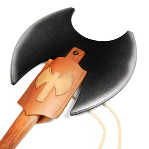 Könnte beinhalten: Eine Doppelaxt mit einem großen, mattschwarzen, halbmondförmigen Kopf an einem Holzstiel. Eine hellbraune Lederscheide bedeckt den unteren Teil des Axtkopfes und ist mit einem aufgenähten, axtförmigen Lederemblem sowie einer hellen Kordel versehen.