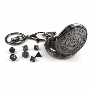Pode incluir: Uma caixa de estilo relógio de bolso em metal cinza escuro com um design de dado de 20 faces na tampa, presa a um porta-chaves. Sete dados poliédricos em miniatura de metal escuro a condizer estão dispostos em frente à caixa aberta.