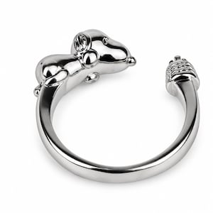 May include: A polished silver-tone open ring featuring a three-dimensional figure of a dog with floppy ears on one end and a textured, layered tail design on the other. The band is smooth and circular, designed to wrap around a finger.
