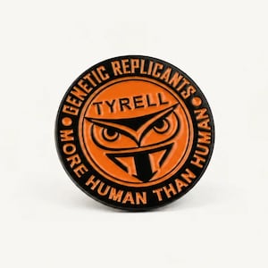 Könnte beinhalten: Ein runder Emaille-Pin in Schwarz und Orange. In der Mitte befindet sich eine Eulen-Grafik über dem Schriftzug TYRELL. Der äußere Rand enthält den Text GENETIC REPLICANTS und MORE HUMAN THAN HUMAN, getrennt durch zwei kleine Punkte.