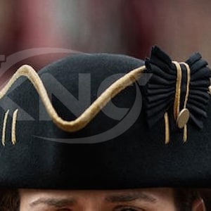 May include: A black tricorn hat with gold trim and accents. The hat features a decorative black bow with gold detailing and a small gold button. The hat's brim is outlined with gold trim.